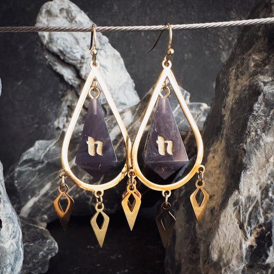 Gold and purple teardrop-shaped dice earrings on a dark stone background